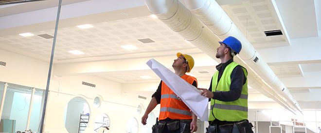 Two workers looking up at the ceiling with blueprints in their hands