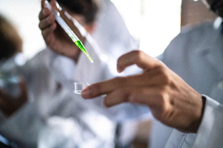 A pair of hands are holding a petri dish while the other hand holds a pipe over the dish.