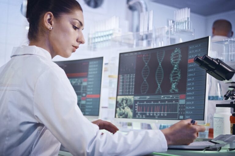 A woman in a white coat sitting at a desk in a laboratory, examining medical charts on her computer