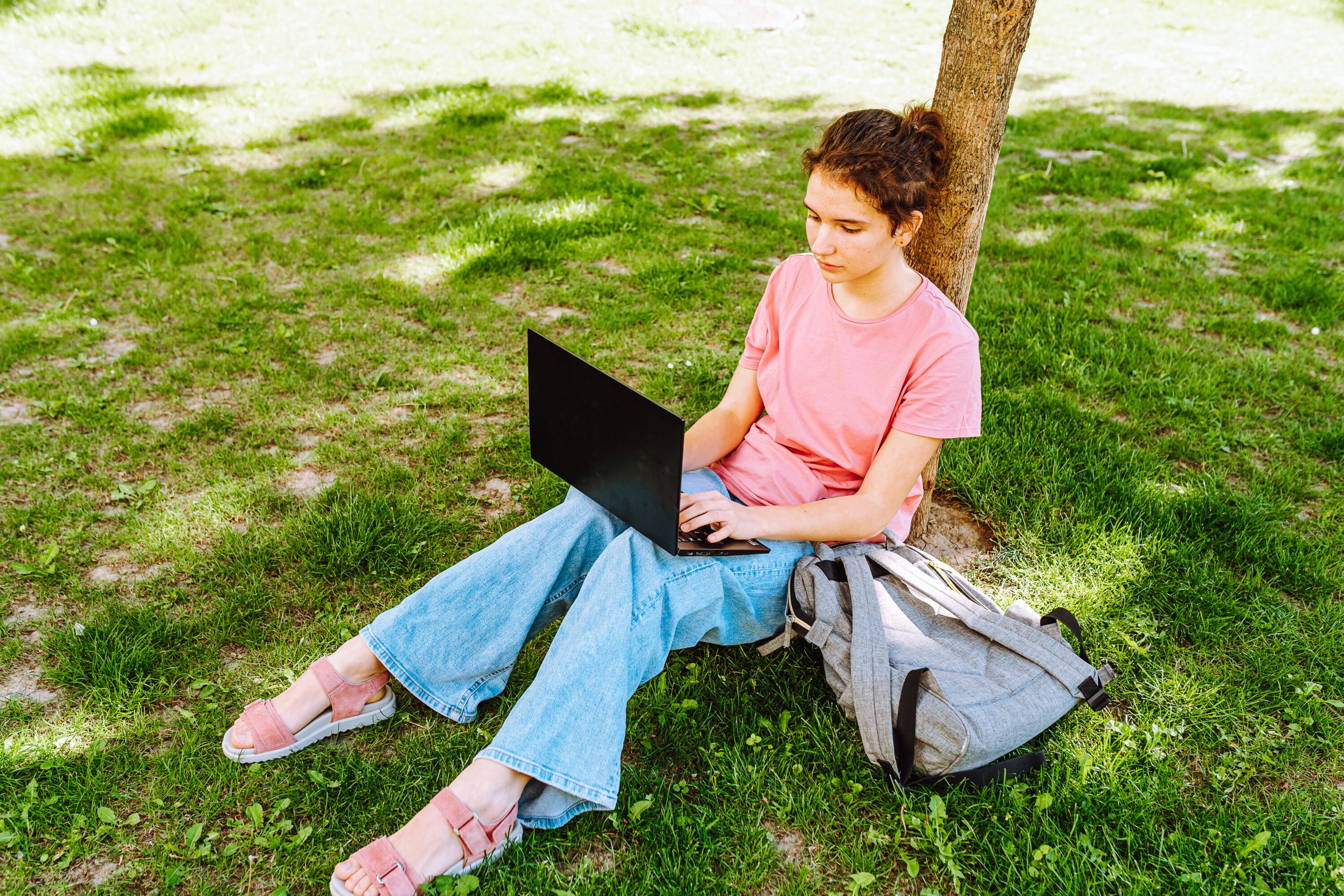 A young woman is on her laptop, sitting on grass, leaning against a tree.