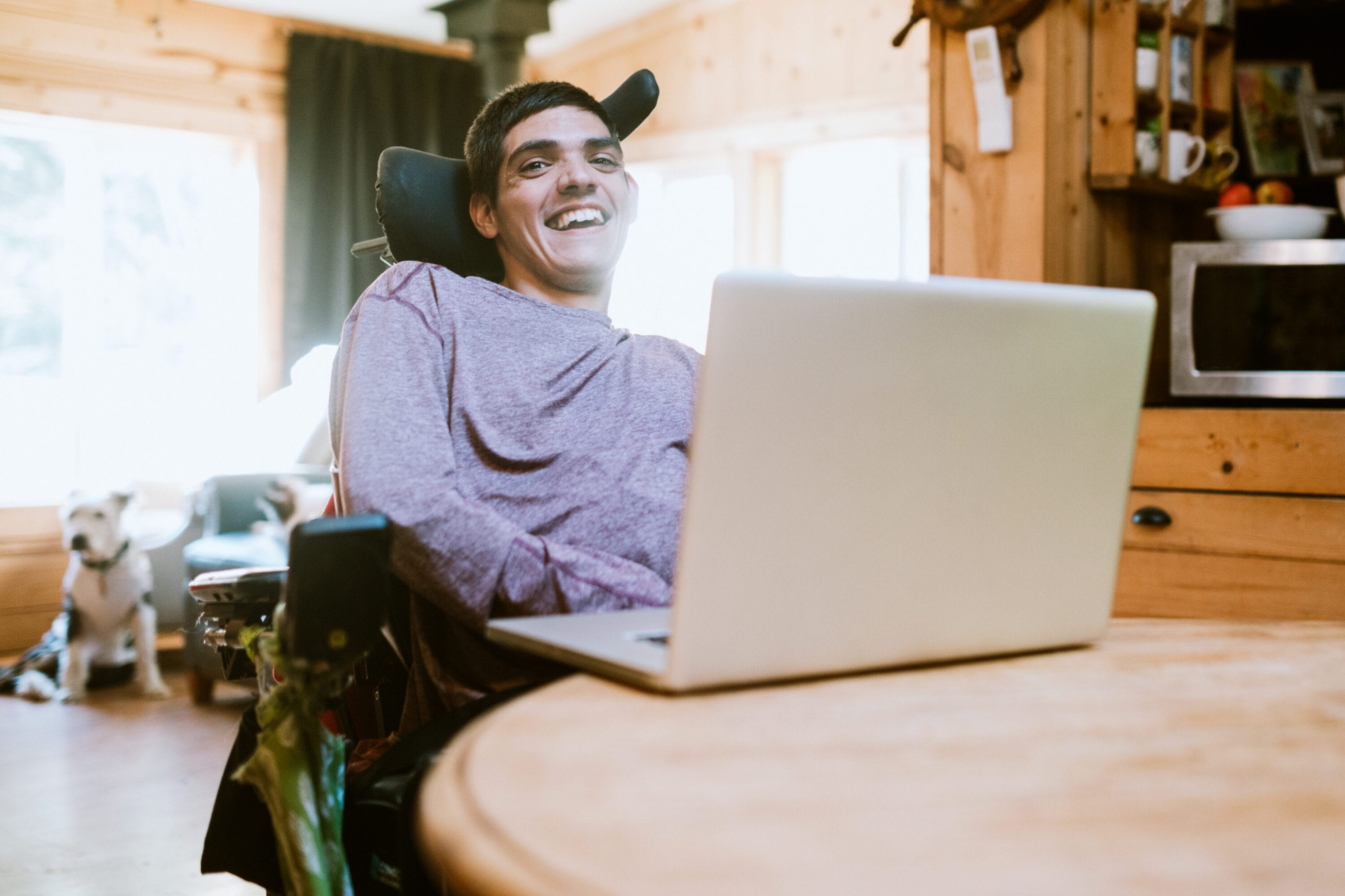 A young man with cerebral palsy is smiling at the camera with a laptop in front of him.