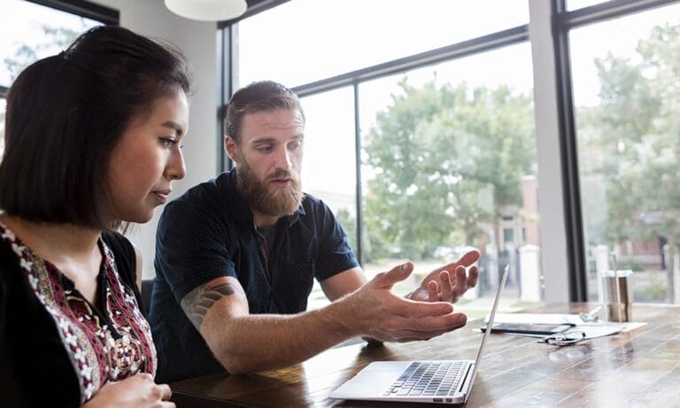 A man and a woman sitting side by side, looking at a laptop in front of them on a table