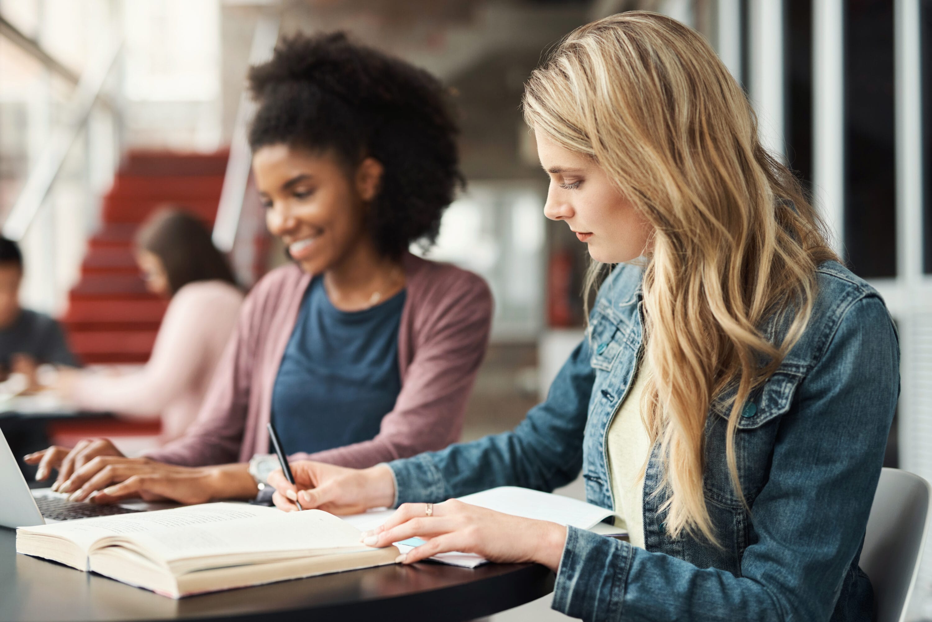 Two women sitting at a table, taking notes with their laptops in front of them.