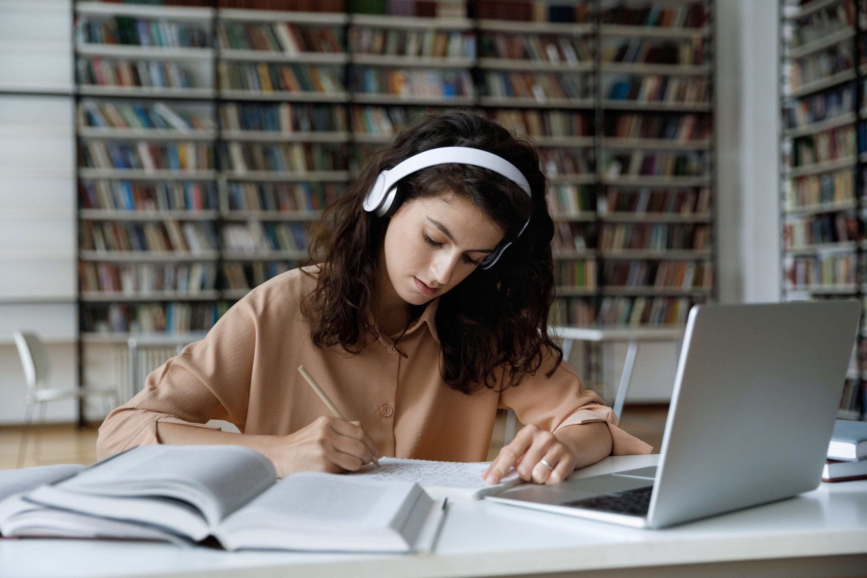 A woman wearing white headphones writing in a notebook in a library.