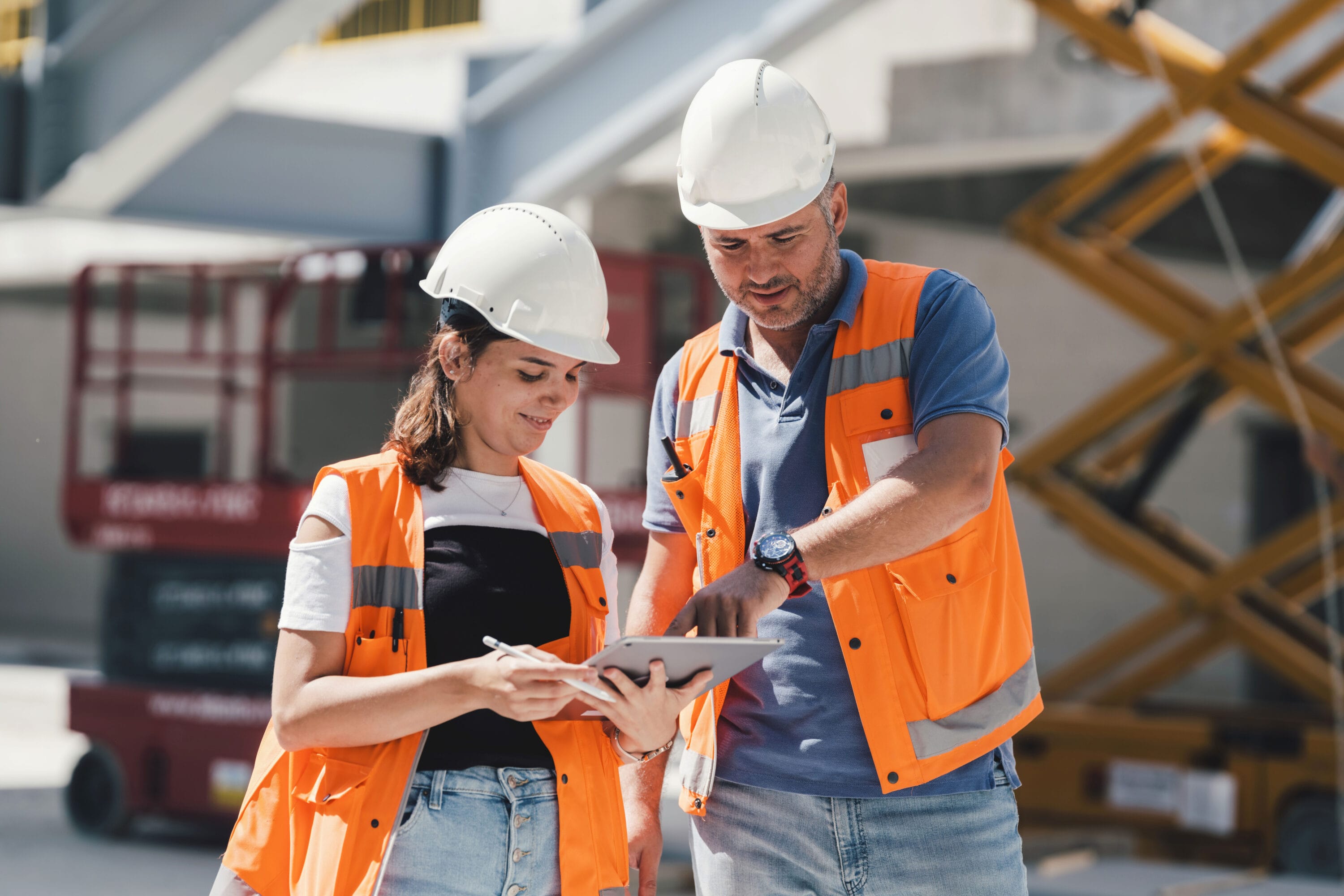 A man and a woman wearing hardhats and orange vests are on-site, reviewing documents on a clipboard.